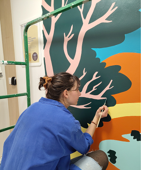 Lise, placée de 3/4 dos à nous, en train de peindre une fresque murale colorée.