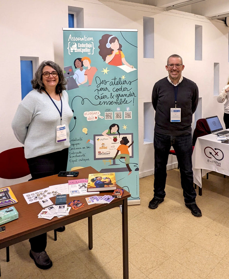 Photo représentant un stand accompagné du roll-up réalisé pour l'association Coder Dojo lors d'une
              journée Unesco et deux bénévoles souriants l'entourant.