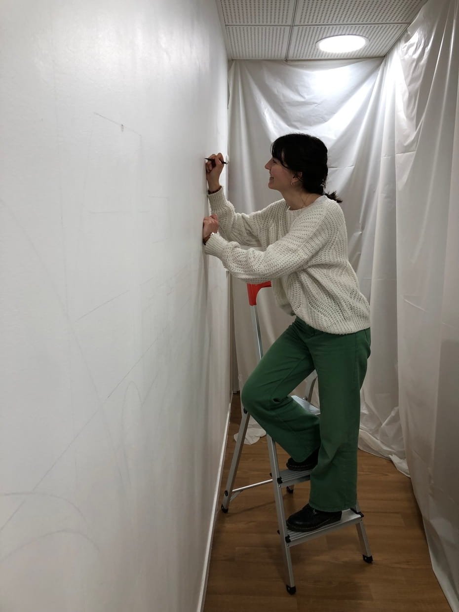 Photo représentant Lise, sur un petit escabeau en train de réaliser l'esquisse préalable de sa fresque murale au crayon à papier.